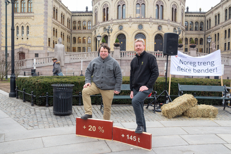 Bjørn Gimming og Tor Jacob Solberg poserer foran Stortinget med foten på avkuttet plankebiter som symboliserer normeringskravet og timene i et årsverk bøndene jobber mer enn andre.